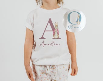 May include: White t-shirt with a pink letter A and a Peter Rabbit illustration. The name Amelia is written below the letter.