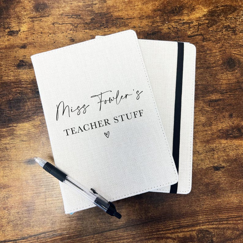 May include: Two cream-coloured notebooks with the text "Miss Fowler's TEACHER STUFF" and a heart symbol. A black and clear pen rests on the notebook. One notebook has a black elastic band. The notebooks are on a wooden surface.