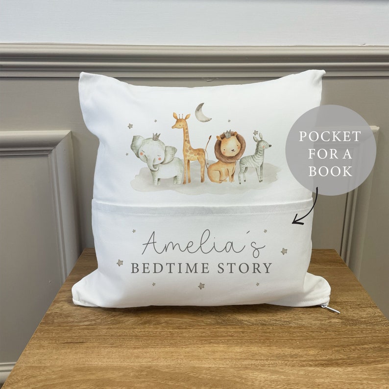 May include: White pillow with a pocket for a book. The pillow has a design of a giraffe, elephant, lion, and zebra with stars and a crescent moon. The text "Amelia's Bedtime Story" is printed on the pillow.