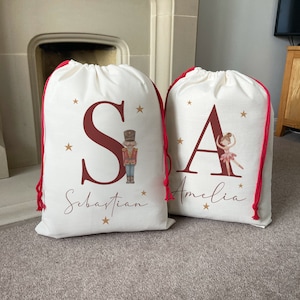 Puede incluir: Dos bolsas de regalo de Navidad personalizadas con cierres de cordón rojo. La bolsa de la izquierda tiene una "S" roja con una ilustración de Cascanueces y el nombre "Sebastian". La bolsa de la derecha tiene una "A" roja con una ilustración de bailarina y el nombre "Amelia".