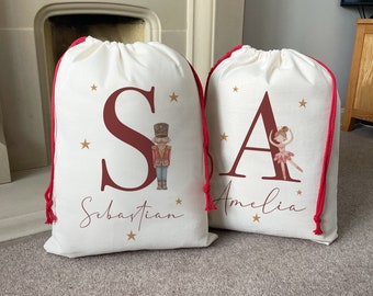 May include: Two personalized Christmas gift bags with red drawstring closures. The bag on the left has a red "S" with a Nutcracker illustration and the name "Sebastian". The bag on the right has a red "A" with a ballerina illustration and the name "Amelia".