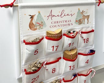 Puede incluir: Calendario de Adviento navideño con bolsillos numerados. La tela blanquecina tiene ribetes rojos y está decorada con ilustraciones de animales del bosque y árboles de Navidad. El texto "Amelia's Christmas Countdown" está impreso en la parte superior.
