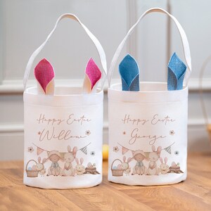 Puede incluir: Dos cestas de Pascua de lona blanca con orejas de conejo. La cesta de la izquierda tiene orejas rosas y dice "Feliz Pascua Willow". La cesta de la derecha tiene orejas azules y dice "Feliz Pascua George". Ambas cestas presentan un lindo diseño de conejito y pollito.