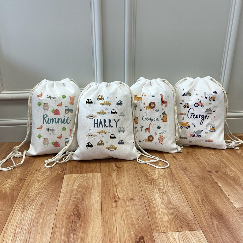 May include: Four white drawstring bags with different designs and names printed on them. The bags feature a farm theme, a car theme, a jungle theme, and a construction theme. The names on the bags are Ronnie, Harry, Jenson, and George.