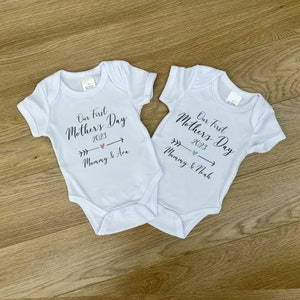 May include: Two light blue baby onesies with the text "Our First Mother's Day 2023" and arrows pointing to the right. One onesie says "Mommy & Ava" and the other says "Mommy & Noah".