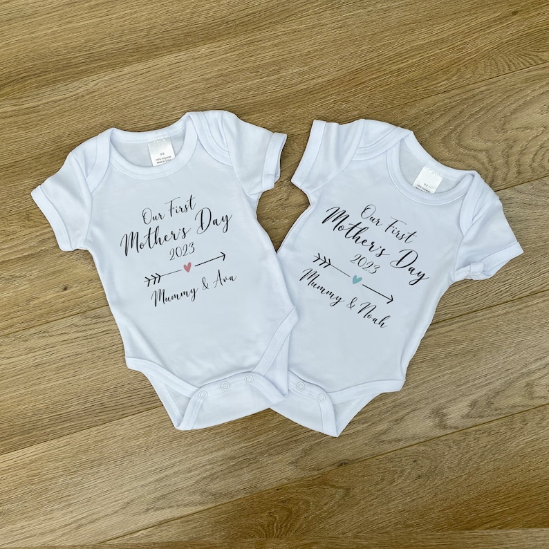May include: Two light blue baby onesies with the text "Our First Mother's Day 2023" and arrows pointing to the right. One onesie says "Mommy & Ava" and the other says "Mommy & Noah".