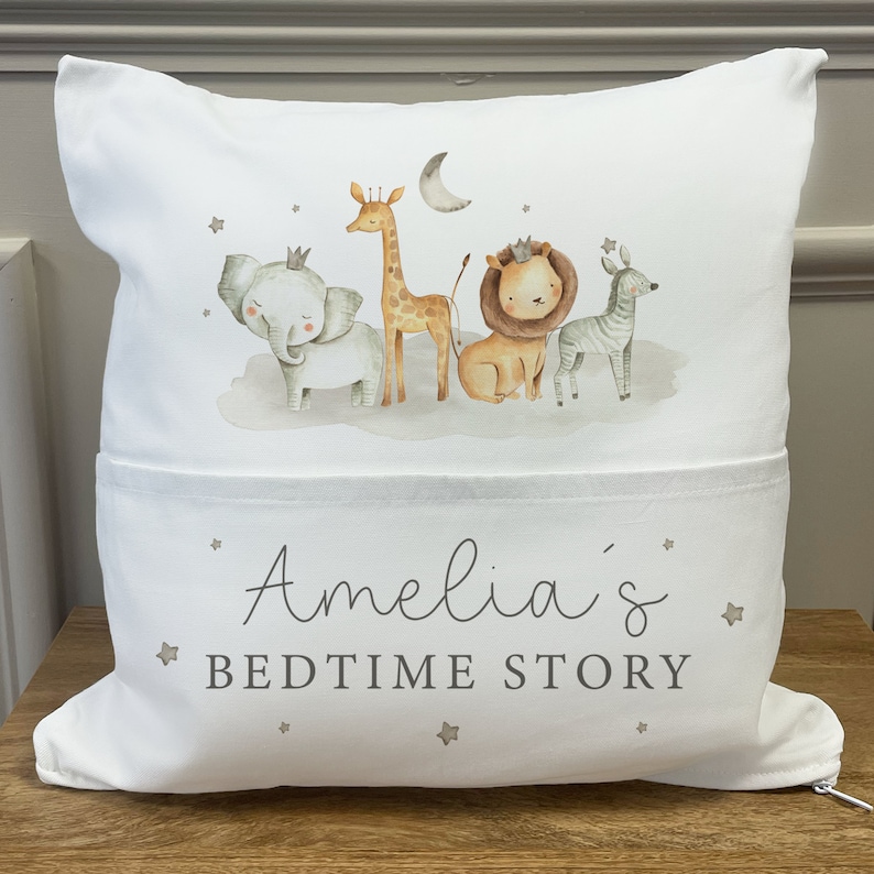 May include: White pillow with a pocket on the bottom. The pillow has a design of a giraffe, elephant, lion, and zebra with a crescent moon and stars. The text "Amelia's Bedtime Story" is printed on the pocket.