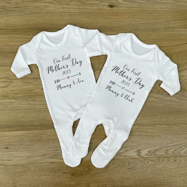 May include: Two white baby onesies with the text "Our First Mother's Day 2023" and arrows pointing to the right. One onesie says "Mommy & Ava" and the other says "Mommy & Noah".