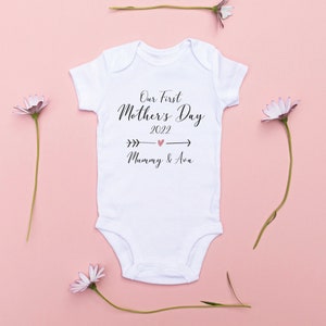 May include: White baby onesie with black text that reads "Our First Mother's Day 2022" with a pink heart and arrows pointing to the left and right. Below the text is "Mummy & Ava" in black text.