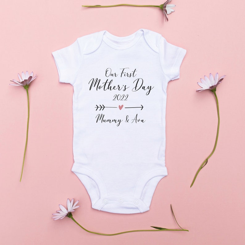 May include: White baby onesie with black text that reads "Our First Mother's Day 2022" with a pink heart and arrows pointing to the left and right. Below the text is "Mummy & Ava" in black text.