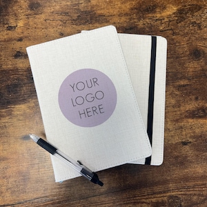 Könnte beinhalten: Zwei cremefarbene Notizbücher mit einem schwarzen Gummiband und einem Stift auf einer Holzoberfläche. Ein Notizbuch hat einen lila Kreis mit dem Text "YOUR LOGO HERE". Der Stift ist schwarz und transparent.