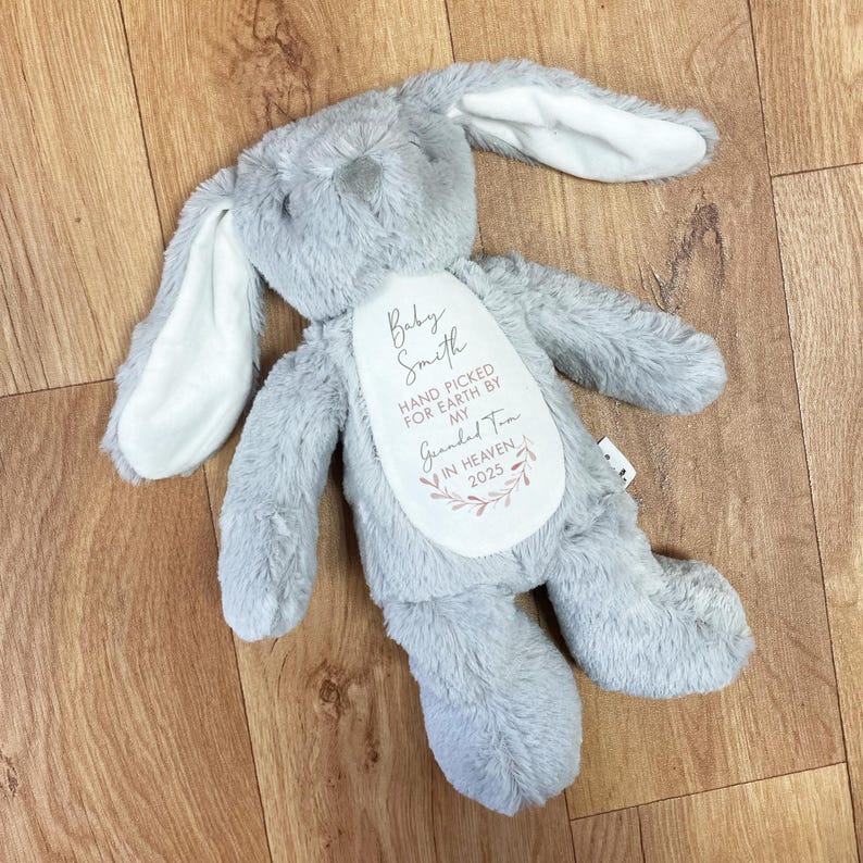 May include: A gray plush bunny with long ears. The bunny has a white patch on its chest with the text "Baby Smith Hand Picked For Earth By My Grandad Tom In Heaven, 2023".