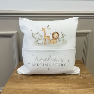 May include: White pillow with a pocket on the front. The pillow has a print of a giraffe, elephant, lion, and zebra with a crown on the elephant and lion. The text "Amelia's Bedtime Story" is printed on the pocket.