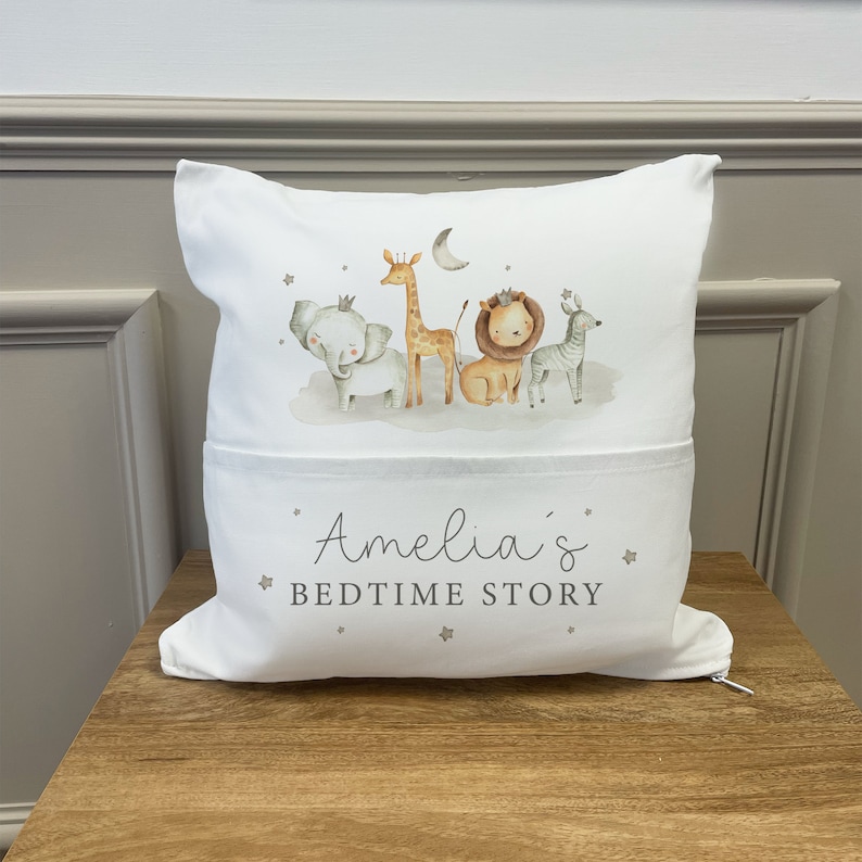 May include: White pillow with a pocket on the front. The pillow has a print of a giraffe, elephant, lion, and zebra with a crown on the elephant and lion. The text "Amelia's Bedtime Story" is printed on the pocket.