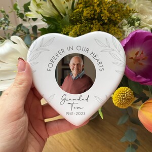 May include: A white heart-shaped memorial plaque with a photo of a man. The plaque has the text "FOREVER IN OUR HEARTS" and "Grandad Tom 1941-2023". Decorative leaf design around the edge. The plaque is held in front of a floral arrangement.