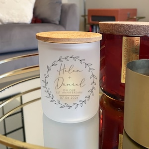 Könnte beinhalten: Ein mattes Glas-Kerzengefäß mit einem Bambusdeckel. Die Kerze hat ein schwarz-weißes Design mit den Namen "Helen + Daniel" und dem Text "On our 1st Anniversary 07.06.2024".