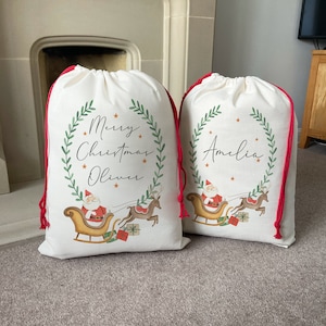 Pu&ograve; includere: Due sacchetti regalo di Natale bianchi con laccetti rossi. I sacchetti presentano una ghirlanda verde con le parole "Buon Natale" e un nome, "Oliver" su un sacchetto e "Amelia" sull'altro. Ogni sacchetto ha un'immagine di cartone animato di Babbo Natale su una slitta trainata da renne.