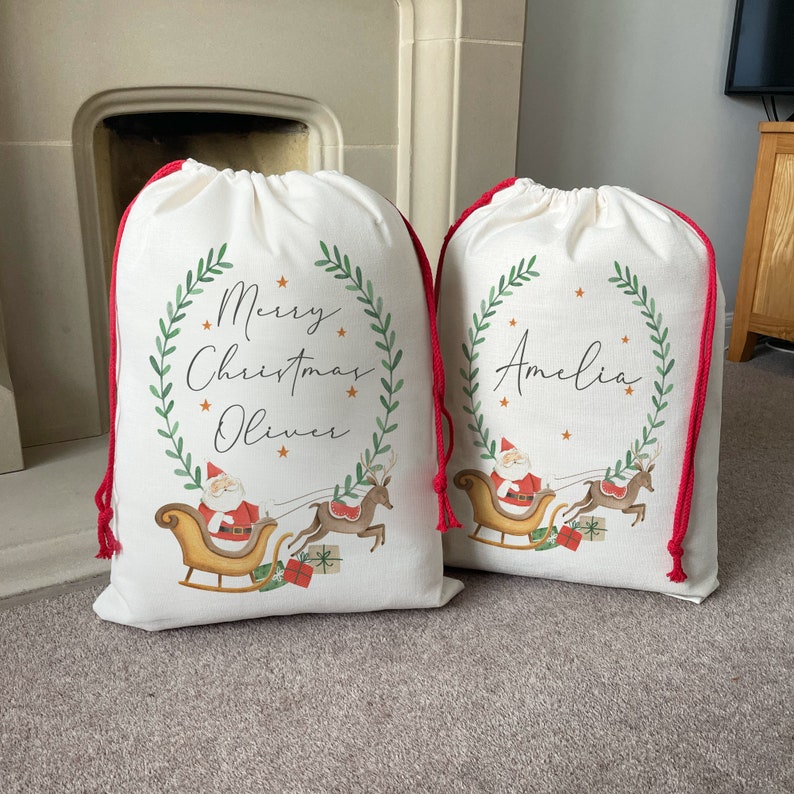 Pu&ograve; includere: Due sacchetti regalo di Natale bianchi con laccetti rossi. I sacchetti presentano una ghirlanda verde con le parole "Buon Natale" e un nome, "Oliver" su un sacchetto e "Amelia" sull'altro. Ogni sacchetto ha un'immagine di cartone animato di Babbo Natale su una slitta trainata da renne.