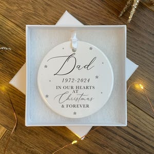 Peut inclure: Ornement de Noël en céramique blanche avec du texte noir qui dit "Dad 1972-2024 In Our Hearts At Christmas & Forever" avec de petites étoiles argentées autour du texte.