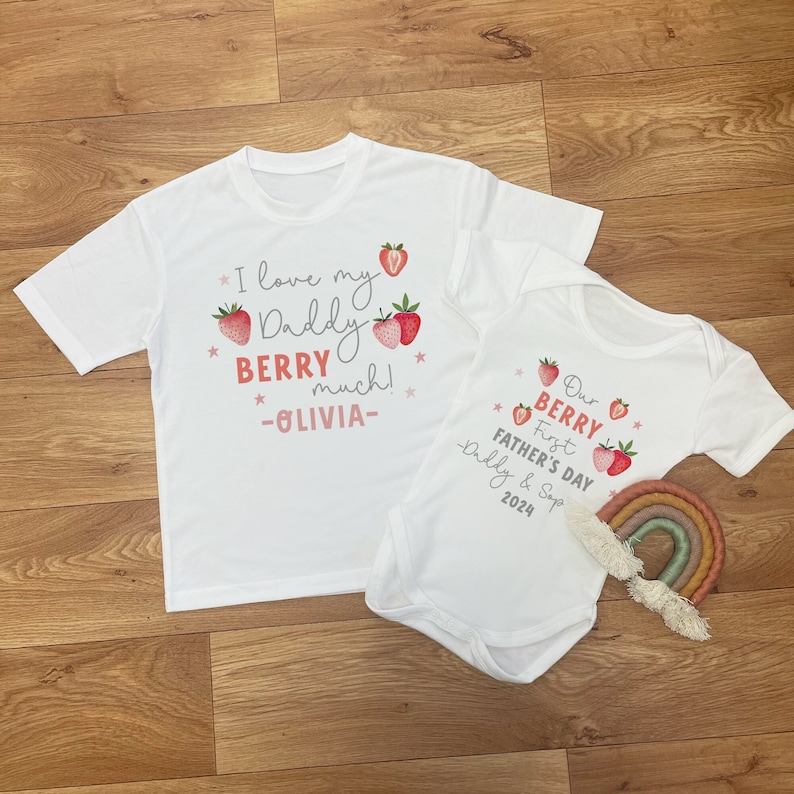 May include: Two white t-shirts with a strawberry theme. The larger shirt says "I love my Daddy BERRY much! -OLIVIA-." The smaller shirt says "Our First BERRY Father's Day -Daddy & Sop 2024."