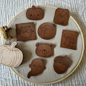 Könnte beinhalten: Eine Sammlung brauner Lederaufnäher mit verschiedenen herbstlichen Motiven, darunter ein Kürbis, ein Fuchs, eine Eule, ein Igel, ein Pilz, ein Bär, ein Geist und eine Eichel. Auch ein Holzkürbis ist vorhanden. Die Aufnäher sind auf beigem Stoff in einem Holzstickrahmen angeordnet.