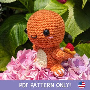 Può includere: Un personaggio di Pokemon all'uncinetto arancione e bianco, Charmander, seduto su un letto di fiori rosa. Il personaggio ha occhi neri e una piccola fiamma rossa sulla coda. L'immagine include il testo "PDF PATTERN ONLY!" e una piccola bandiera americana.