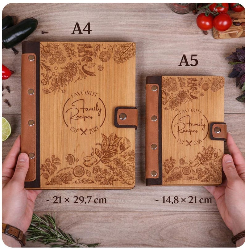 May include: Two wooden recipe books, A4 and A5 sizes, with brown leather accents. The covers are engraved with botanical designs and the text "FAVORITE Family Recipes EST X 2021". The A4 book measures approximately 8.3 x 11.7 inches, and the A5 book is about 5.8 x 8.3 inches.