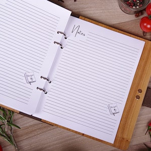 May include: Open wooden recipe journal with lined pages and the word "Notes" in script. The journal has a leather strap and metal rings. Small illustrations of recipe books are on the pages. Fresh tomatoes, herbs, and spices surround the journal.