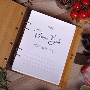 May include: A wooden recipe book with the text "This Recipe Book Belongs To:" on the open page. The book has a light brown wooden cover and a dark brown leather spine. The book is open on a wooden surface with tomatoes and herbs.