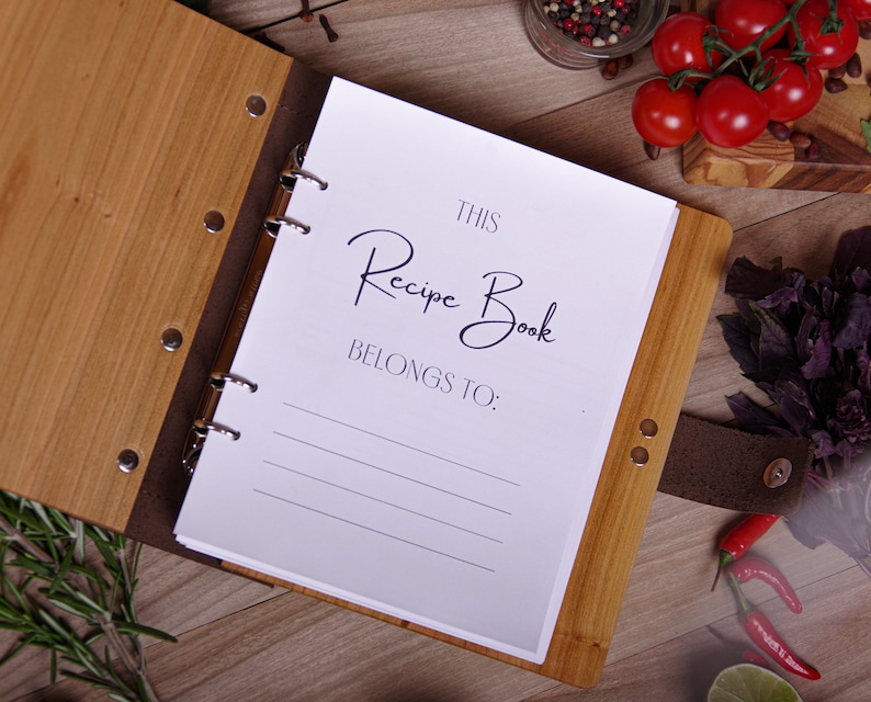 May include: A wooden recipe book with the text "This Recipe Book Belongs To:" on the open page. The book has a light brown wooden cover and a dark brown leather spine. The book is open on a wooden surface with tomatoes and herbs.