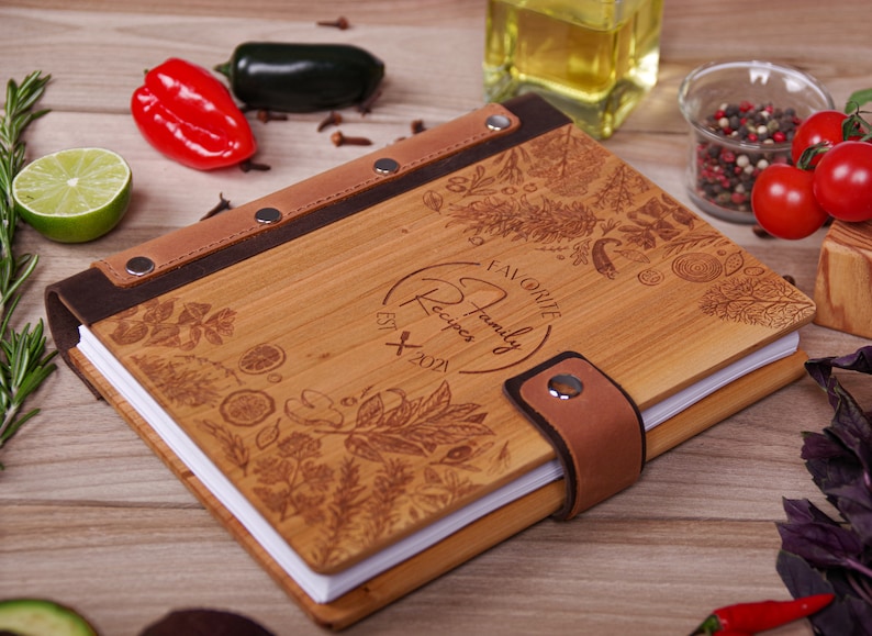 May include: A wooden recipe book with a brown leather spine and strap closure. The cover is engraved with floral and herb designs, and the text "Favorite Family Recipes EST 2021". The book is open, revealing white pages.