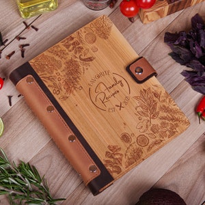 May include: Wooden recipe book with a light brown cover and dark brown leather spine and accents. The cover is engraved with botanical illustrations and the text "Favorite Family Recipes Est. 2021". A leather strap with a snap closure secures the book.