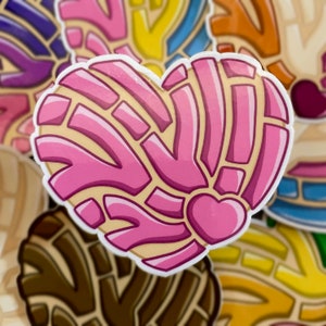 May include: A heart-shaped sticker featuring a pink concha bread design with a textured, segmented topping pattern. The center of the heart includes a smaller, solid pink heart shape, all outlined in white against a background of other stickers.