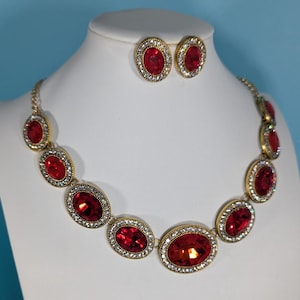 May include: A gold-coloured necklace and matching earrings set. The necklace and earrings feature large, oval, red gemstones surrounded by a halo of small, clear crystals. The jewellery is displayed on a white mannequin head against a blue background.