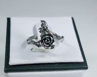 Könnte beinhalten: Silberring mit Rosenmotiv. Die Rose befindet sich in der Mitte des Rings und ist von Blättern umgeben. Der Ring liegt auf einer weißen Schachtel mit schwarzem Rand.
