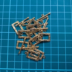 Op de afbeelding: Een stapel goudkleurige metalen frameversieringen met sierlijke details. De frames zijn rechthoekig en hebben een kleine lus aan de bovenkant. De versieringen zijn verspreid over een groene snijmat.