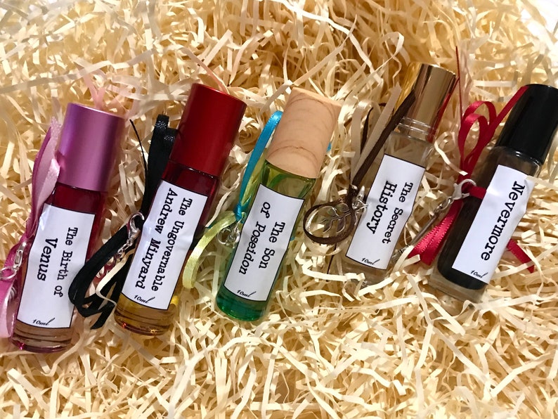May include: Six small glass bottles with roll-on applicators, each containing a different scented oil. The bottles are labeled with the names of books: "The Ungovernable, Andrew Minyard", "The Son of Poseidon", "The Secret History", "Nevermore", and "The Birth of Venus".