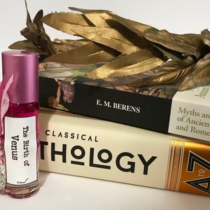 May include: A pink roll-on bottle with a white label that reads "The Birth of Venus". The bottle is sitting on top of two books. The top book is black with white text that reads "E.M. Berens" and "Myths and Legends of Ancient Greece and Rome". The bottom book is cream colored with black text that reads "CLASSICAL THOLOGY" and has a gold border with a design on the right side that reads "A to Z".