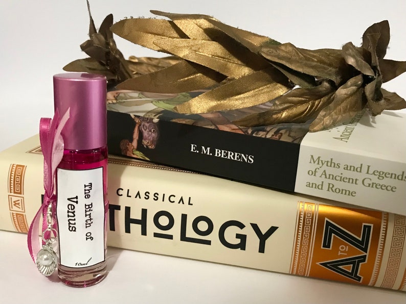 May include: A pink roll-on bottle with a white label that reads "The Birth of Venus". The bottle is sitting on top of two books. The top book is black with white text that reads "E.M. Berens" and "Myths and Legends of Ancient Greece and Rome". The bottom book is cream colored with black text that reads "CLASSICAL THOLOGY" and has a gold border with a design on the right side that reads "A to Z".