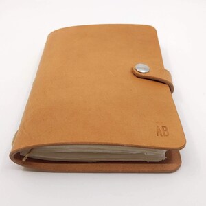 May include: A tan leather bound journal with a silver button closure and the initials "AB" embossed on the front cover.
