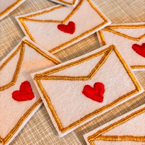 Può includere: Toppe a forma di busta in feltro bianco con bordi dorati e un cuore rosso. Le toppe sono disposte su una superficie strutturata. Un affascinante oggetto artigianale.
