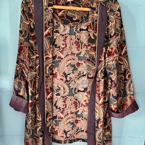 May include: A vintage paisley print robe in shades of burgundy, green, and tan. The robe features a dark purple trim and is hanging on a wooden hanger. The robe has a classic, elegant design.