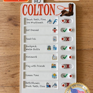 May include: A white plastic chore chart with the name COLTON printed in orange at the top. It features eight rows of tasks with sliding toggles to mark completion with a green checkmark or red X. Includes icons for daily activities and a cowboy boot.