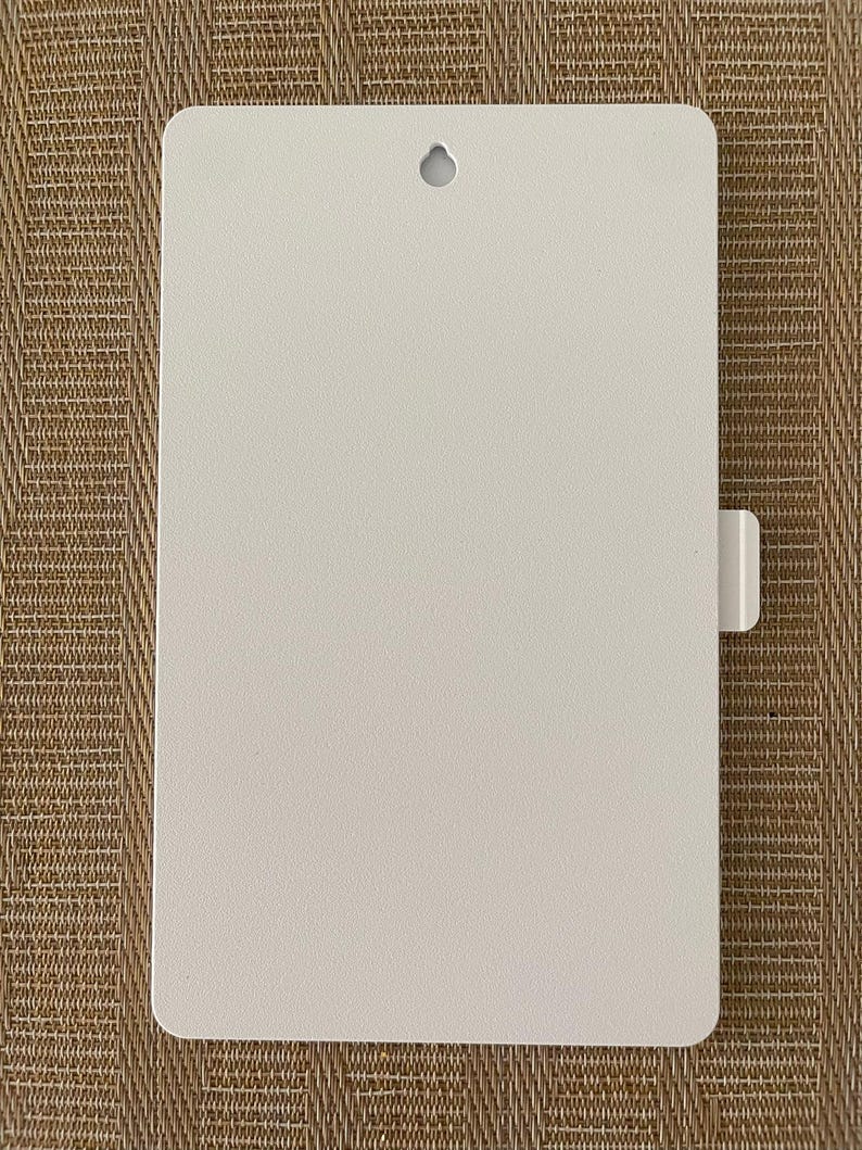 May include: A rectangular, off-white metal sign with rounded corners. It features a small, circular hole at the top for hanging and a rectangular tab on the right side. The sign has a textured surface.
