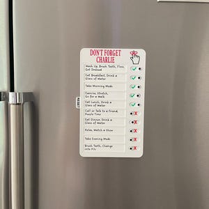 May include: A white magnetic checklist with the text "DON'T FORGET CHARLIE" and daily tasks. The checklist includes tasks like "Wash Up, Brush Teeth, Floss, Get Dressed" and "Take Morning Meds". Check marks and X's indicate completed tasks.