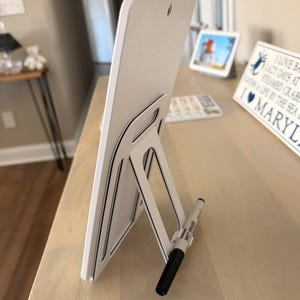 May include: White rectangular sign with a black and white stand and pen holder. The sign has a textured surface and a small hole at the top. A black and white wet erase marker is in the pen holder. Text on the sign reads "Optional Stand & Pen Holder."