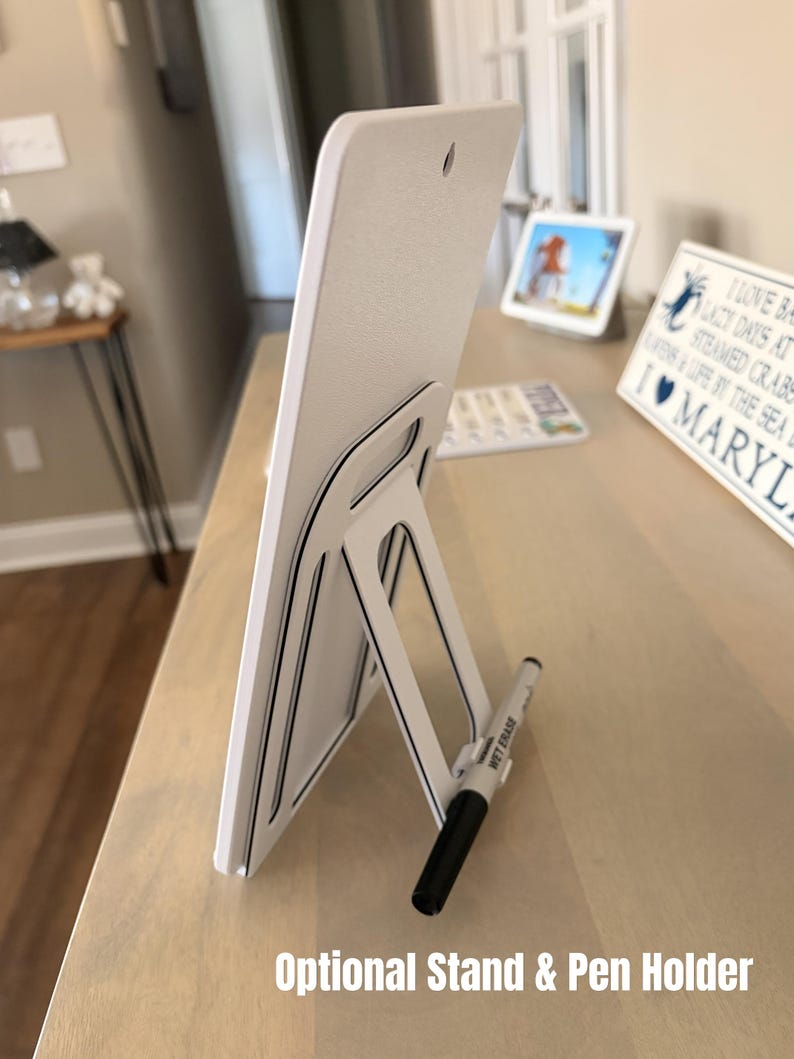 May include: White rectangular sign with a black and white stand and pen holder. The sign has a textured surface and a small hole at the top. A black and white wet erase marker is in the pen holder. Text on the sign reads "Optional Stand & Pen Holder."