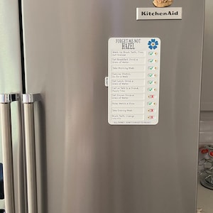 May include: A stainless steel refrigerator with a magnetic checklist that reads "FORGET-ME-NOT HAZEL". The checklist includes daily tasks with checkmarks and X's. A KitchenAid magnet and a lighthouse magnet are also on the refrigerator.