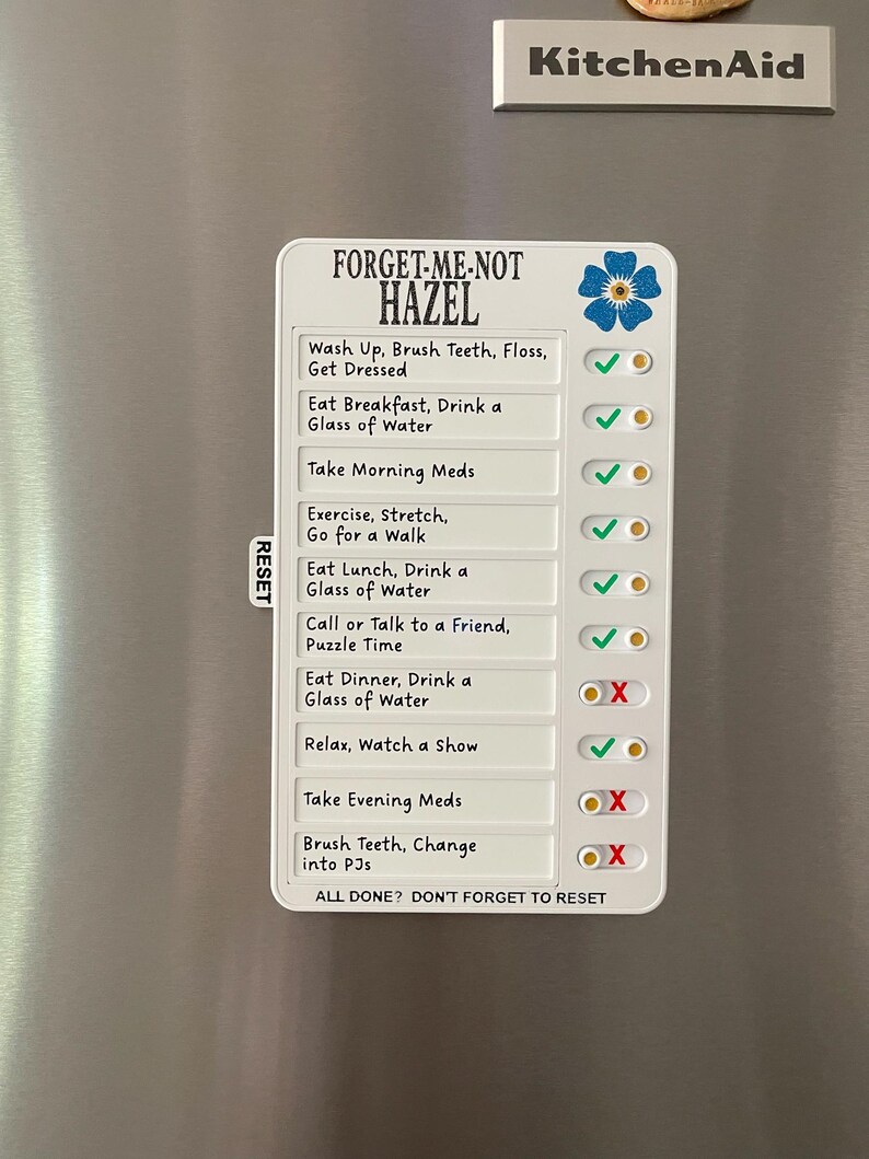 May include: A white magnetic checklist with the text "FORGET-ME-NOT HAZEL" and daily tasks. The checklist includes a blue flower graphic and check boxes with green check marks and red X's. The checklist is on a silver refrigerator.