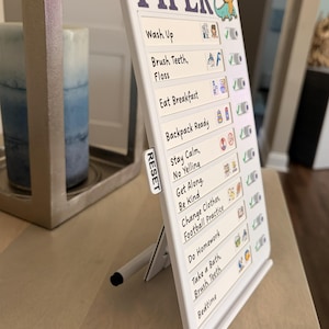 May include: White chore chart with the name "PIPER" in purple. The chart includes a list of tasks with check boxes and a small dinosaur graphic. The chart is propped up by a white stand. The text "Shown With Optional Stand" is at the bottom.